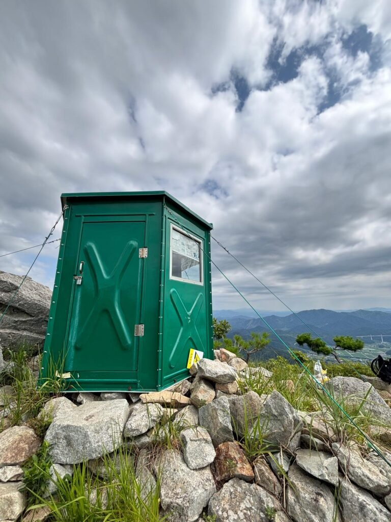 A summit scene on Misungsan, stands an old green hut secured with guy lines, set against cloudy skies and forested hills in the distance.