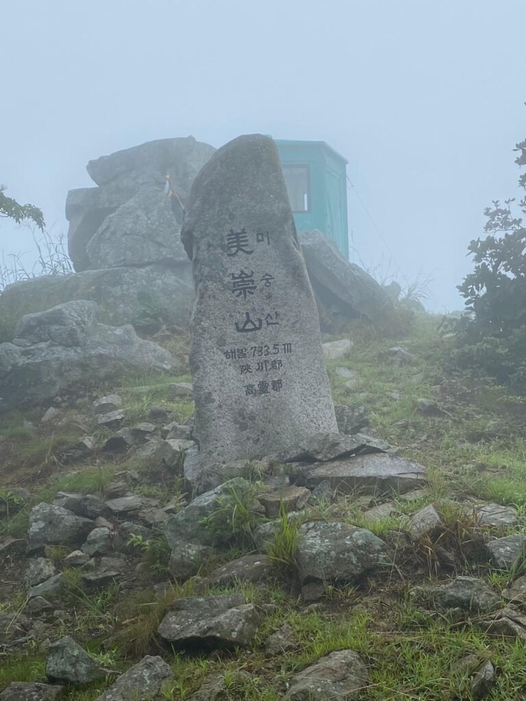 On the summit of Misungsan: a tall stone marker stands in the foreground among rocks, with an old green hut behind it. Low cloud makes the scene misty, and the surrounding landscape is hidden from view.