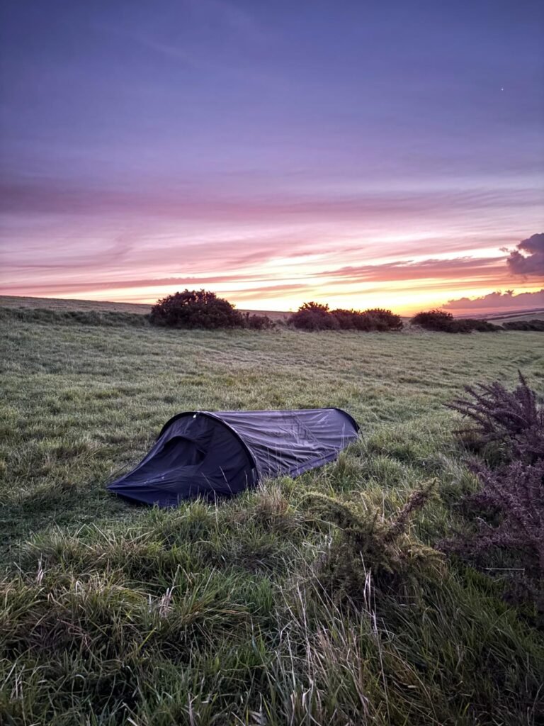 A bivi, on the top of a grassy hill, with the blue, purple and orange sky of dawn above.