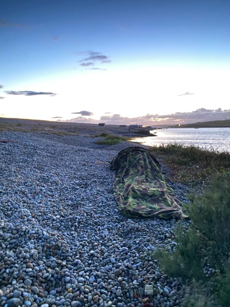 A Dutch hooped bivvi, set up on the pebbled coast next to a patch of short grass and the water.