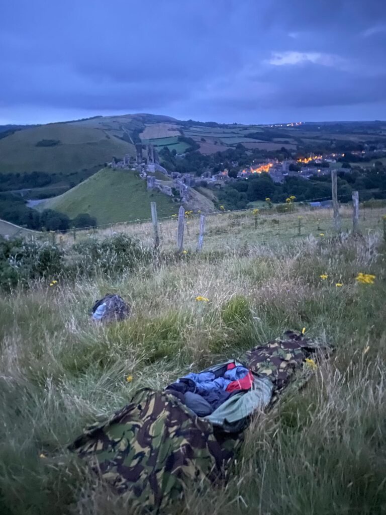 Dutch hooped bivi, open and stuffed with a sleeping bag, on the top of a hill in long grass, and over looking a castle ruins on top of a hill and next to a village.