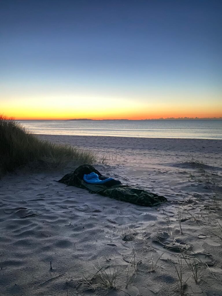 a bivi set up on the beach, surrounded by beach grass, and with the dark blue, and orange sky of dawn on the horizon