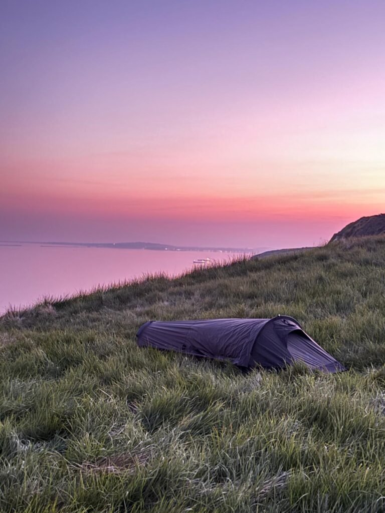 A bivi bag, in grass on top of a hill and overlooking to sea and the purple dusk sky