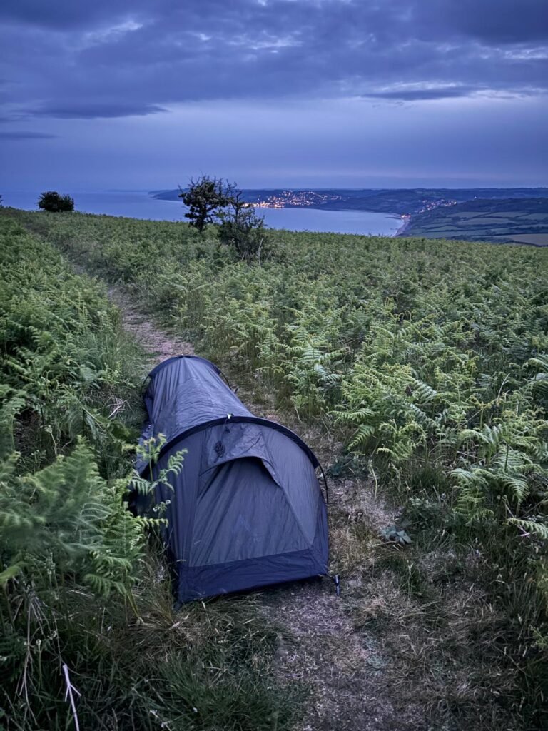 On top of a hill, a Nortent Skjul bivi is set up on a narrow path through ferns, with the coastline visible in the background below, and the illuminated lights of a small coastal town.