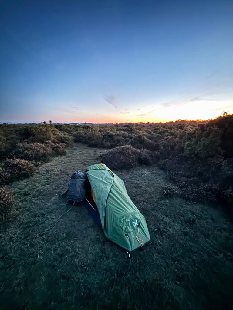 The Phoxx 1v2 tent set up with the side flap open, a backpack leaning against the side of the tent, and a clear sky above and an bright orange horizon of dusk behind.