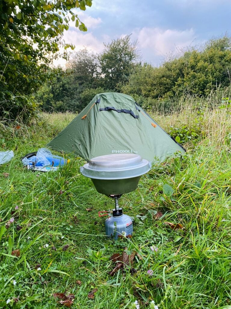 Am image of the OEX Phoxx 1v2 set set up in long grass, with a camping stone and a Trangia cooking pot sitting on top of it, on the ground in front of the tent.