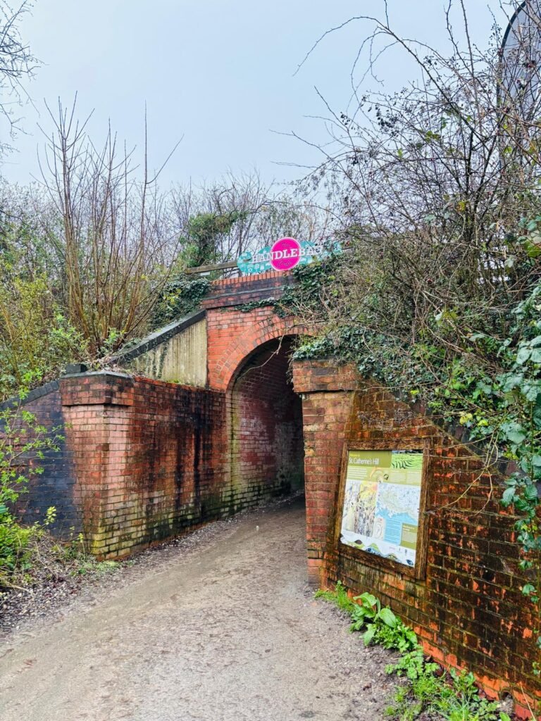 The brick bridge at the Cadence Clubhouse in Winchester, with a map and information board attached to one wall. marks  one of the entrances to St Catherine's Hill in Winchester