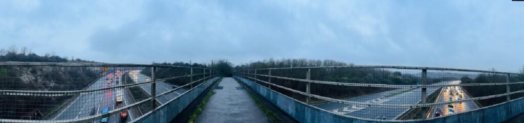 A panoramic photo of the bridge leading over the M3 motorway at St Catherine's Hill, with cars visible on the carriageway below.