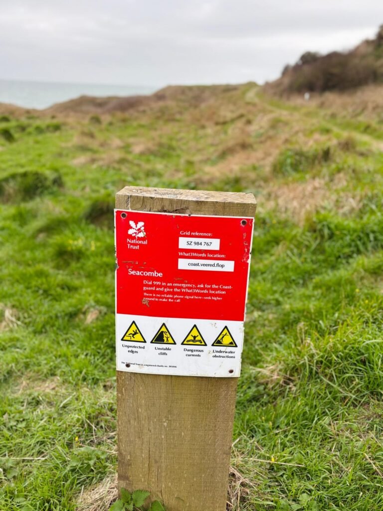 Red warning sign on a wooden post at Seacombe cliff warning of unstable edges, dangerous currents, and underwater hazards