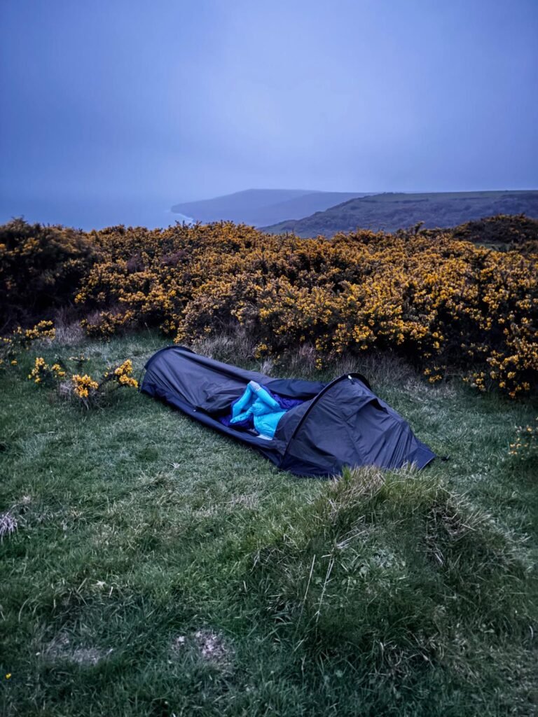 Nortent Skjul bivvy set up on grass, sheltered by gorse bushes with Dorset coastal hills and sea in the distance