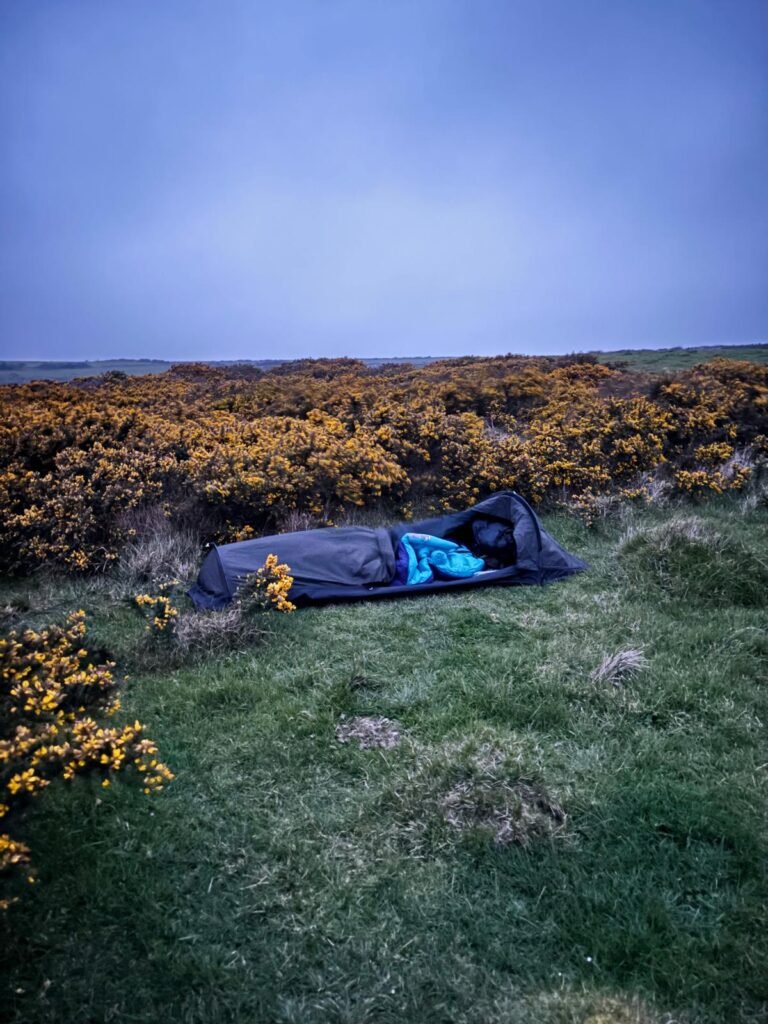 Side view of Nortent Skjul bivvy set up behind gorse bushes with Alpkit sleeping bag visible, low-profile wild camping setup