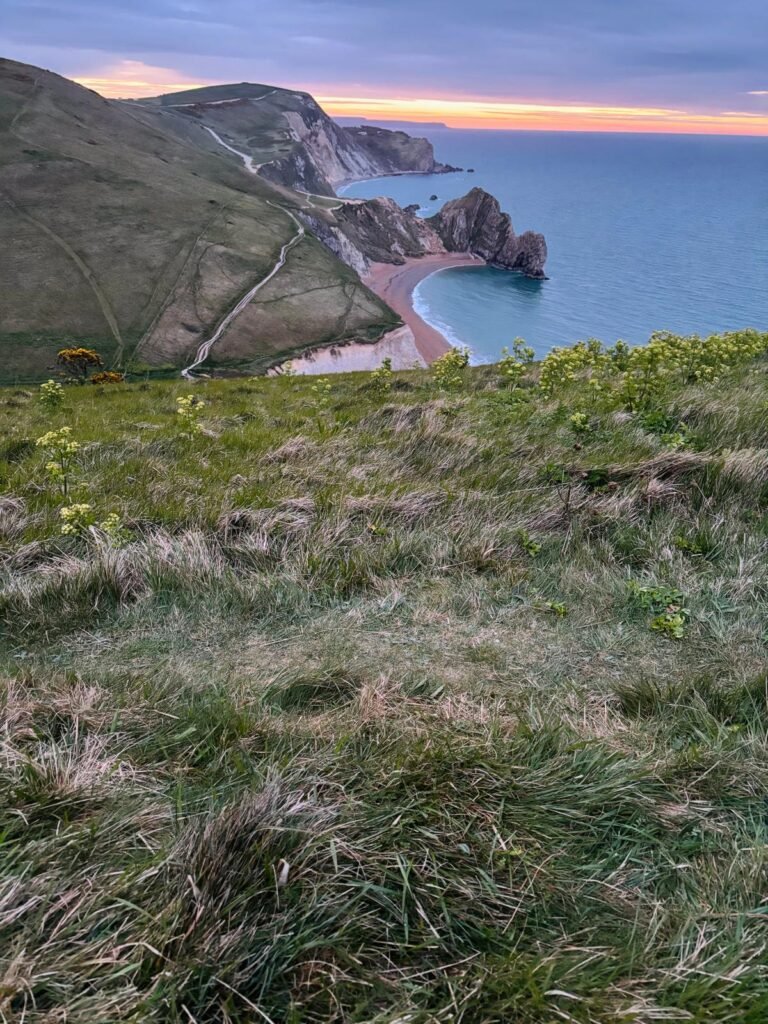 View from Swyre Head looking down at Durdle Door at dawn, with a reddish-orange horizon, clear beach, and the South West Coast Path visible over the hills