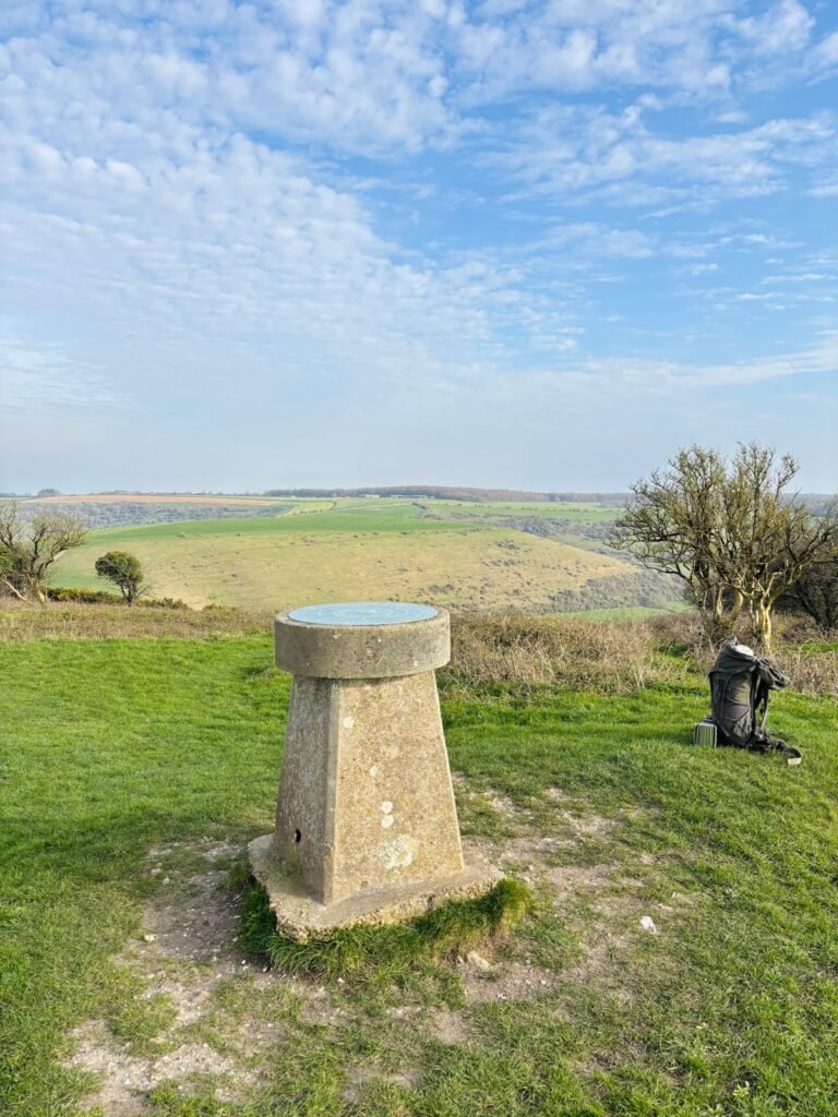 Trig point TP4764 on Melbury Hill with backpack and Dorset hills in the background
