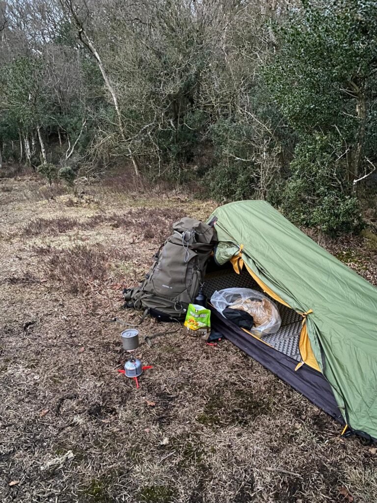 OEX Phoxx 1 V2 tent pitched beside woodland with backpack, stove and sleeping setup during wild camp