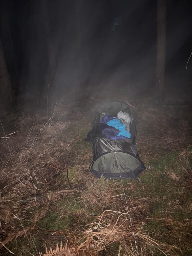 Open bivvi in rain and mist at night in woodland with sleeping bag visible inside.