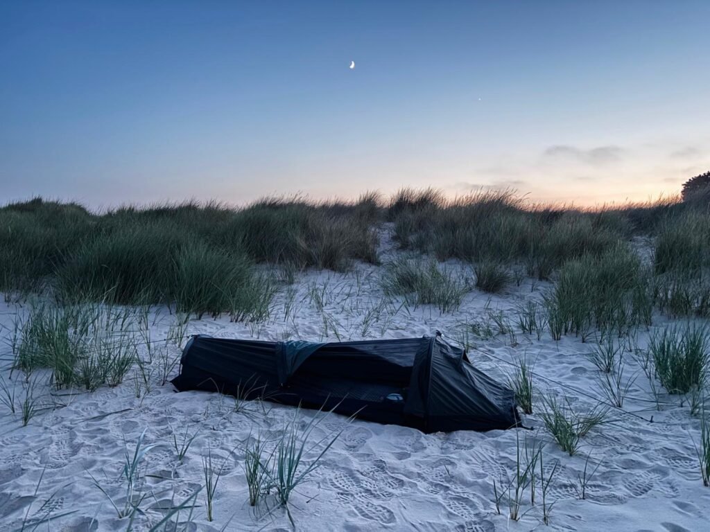 Nortent Skjul bivvy tucked amongst beach grass at dusk, with the moon and a bright planet visible above the orange horizon.