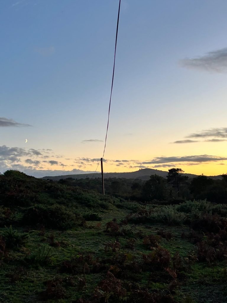 A crescent moon above dark heathland and gorse beneath an orange horizon
